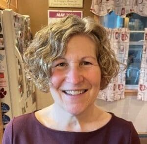 A woman with short, curly gray hair and a maroon top smiles in a kitchen with patterned curtains and a calendar on the wall.