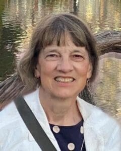 An older woman with short gray hair and a white jacket smiles in front of a body of water with tree reflections.