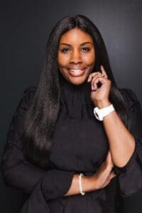 A woman with long straight hair, wearing a black top and white watch, stands against a dark background, smiling and touching her face with one hand.