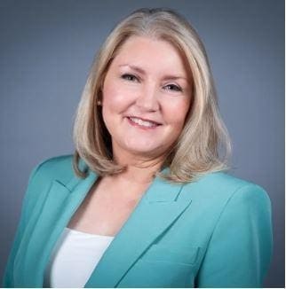 A woman with shoulder-length blonde hair wearing a teal blazer and white top, smiling in front of a plain grey background.