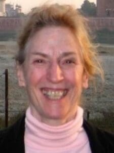 A woman with blonde hair smiles at the camera, wearing a light pink turtleneck and a dark jacket, with an outdoor background visible behind her.