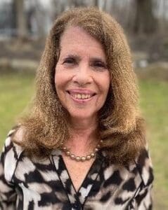 A woman with shoulder-length curly hair, wearing a patterned blouse and a pearl necklace, smiles outdoors with grass and trees in the background.