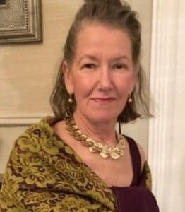 An older woman with gray hair wears a patterned shawl, a gold necklace, and earrings, standing indoors with a slight smile.