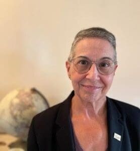 An older person with short gray hair, glasses, and a blazer stands indoors next to a globe, facing the camera and smiling.