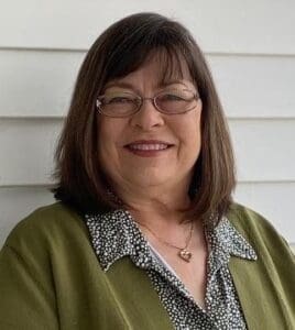 A woman with shoulder-length brown hair, glasses, and a green cardigan smiles at the camera, standing in front of a white paneled wall.