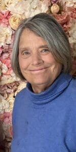 An older woman with gray hair and a blue sweater smiles in front of a background of pink and white flowers.