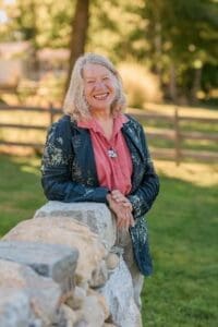 An older woman with gray hair stands outdoors by a stone wall, smiling, wearing a dark jacket and pink blouse. A wooden fence and trees are visible in the background.