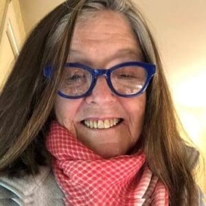 A woman with long brown hair and blue glasses smiles, wearing a red and white checkered scarf and a light-colored jacket.