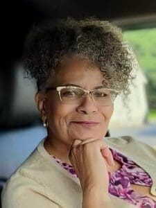 A woman with curly gray hair and glasses, wearing a beige cardigan and purple blouse, sits with her hand resting on her chin, smiling slightly.