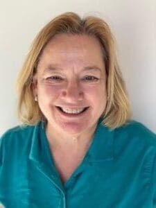 A middle-aged woman with blonde hair, smiling, wearing a teal collared shirt, in front of a plain light-colored background.