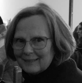 An older woman with glasses and shoulder-length hair smiles at the camera in a black and white photo. Other people are visible in the blurred background.