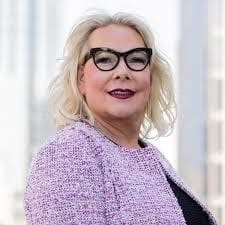 A woman with blonde hair, wearing black cat-eye glasses, a pink textured jacket, and dark lipstick, stands in front of a blurred urban background.