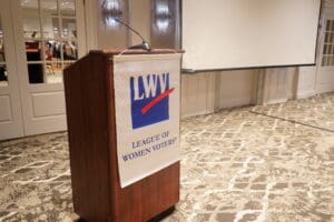 A podium with a League of Women Voters banner stands in a carpeted conference room next to a blank projection screen.