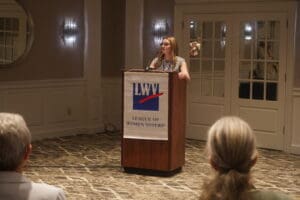 A woman stands at a podium with a "League of Women Voters" sign, speaking to an audience in a conference room.