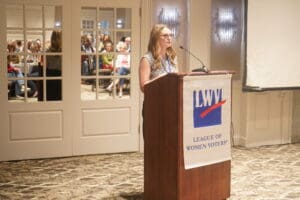 A woman speaks at a podium with a “League of Women Voters” banner in front of an audience in a conference room.