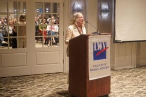 A person speaks at a podium with a "League of Women Voters" banner in a conference room; an audience is visible in the background.