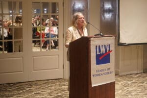 A woman speaks at a podium with an LWV League of Women Voters banner in front of an audience seated in a conference room.