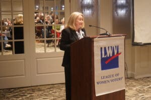 A woman speaks at a podium with a League of Women Voters banner in a conference room, with an audience visible in the background.