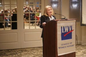 A woman speaks at a podium with a League of Women Voters banner in a conference room; audience members are visible in the background.