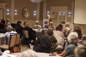 A woman speaks at a podium with an LWV banner to a seated audience in a conference room with round tables and beige walls.