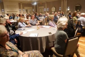 A group of adults sits at round tables in a conference room, attentively listening to a presentation, with notebooks and folders on the tables.