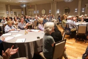A group of people sit at round tables in a banquet hall, listening and applauding during an indoor event or presentation.