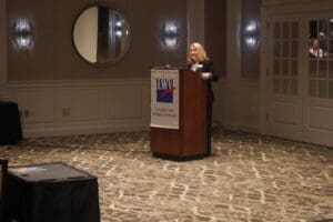 A woman stands at a podium labeled "League of Women Voters," speaking in a conference room with a carpeted floor and wall mirrors.