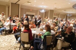 A large group of people, mostly women, sit and stand in a conference room, attentively watching a presentation and clapping. Tables, chairs, and notepads are visible.