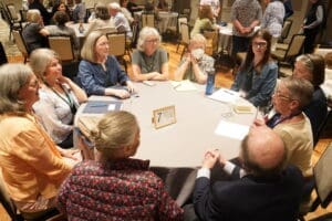 A group of people sit around a round table engaged in discussion at a conference or meeting in a large room.