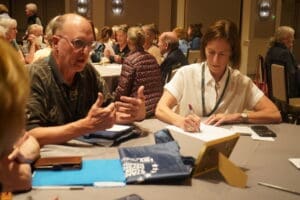 A man speaks while gesturing with his hands as a woman takes notes at a round table during a group meeting or workshop.