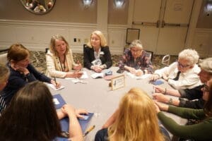 A group of people sit around a round table in a conference room, engaged in discussion and taking notes.