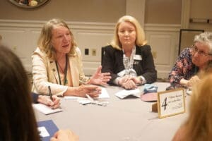 Four women sit around a table engaged in discussion, with notepads and papers in front of them. A sign on the table reads, “4 Where humans and online lives…”.