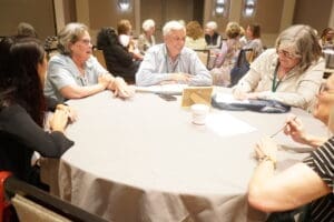 A group of adults sit around a round table engaged in conversation at a conference or meeting room.