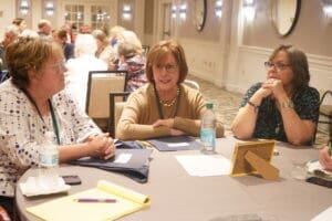 Three women are seated at a round table engaged in conversation, with notepads, folders, and water bottles in front of them in a conference room setting.