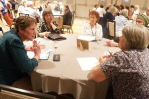 A group of women sit around a table engaged in discussion at a conference or meeting, with notebooks, paper, and glasses of water in front of them.