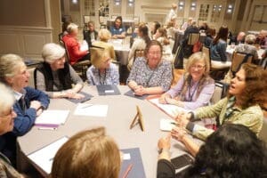 A group of women sit around a round table engaged in discussion, with other groups visible in the background in a well-lit conference room.