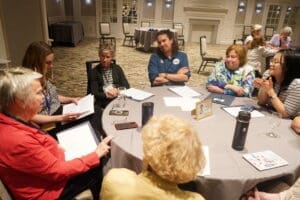 Seven women sit around a round table in a conference room, engaged in discussion with papers and drinks in front of them.