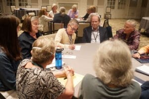A group of older adults sit around a table engaged in discussion at a meeting or workshop in a conference room.