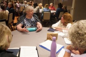 A group of women sit around a round table engaged in discussion, with notebooks, water bottles, and papers in front of them at a conference or meeting.