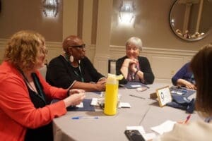 Four women sit around a round table in a conference room, engaged in conversation with notebooks, pens, and a yellow water bottle on the table.