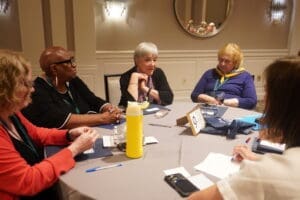 Five women sit around a round table in discussion at a conference, with notebooks, pens, and a yellow water bottle on the table.