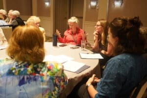 A group of people sit around a round table in discussion, with papers and drinks in front of them, in a conference room setting.