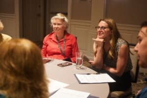 Two women sit at a round table in a meeting room, listening and smiling, with papers, a pen, and a glass of water in front of them.