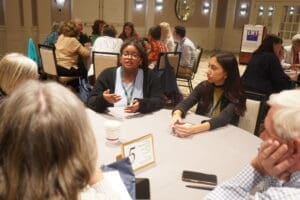 A group of people sit around a table having a discussion at an indoor event, with notepads, a coffee cup, and a table number sign visible.