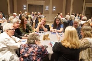 A group of women sit around a table engaged in conversation at a large gathering or conference, with other groups in the background.