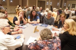 A group of women sit around a table in discussion at a conference or meeting, with papers, notebooks, and phones in front of them.