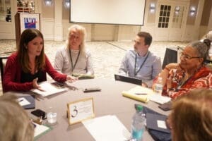 Four people sit around a round table in discussion, with papers, notebooks, and drinks in front of them at a conference or meeting.