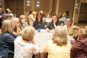 A group of women sit around a round table in discussion at a conference or meeting, with more groups in the background.