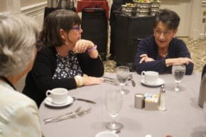 Three women sit at a round table set for a meal, engaged in conversation with water glasses, coffee cups, and utensils in front of them.