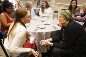 Several women of different ages sit around a round table in conversation at what appears to be a conference or meeting room, with coffee cups and utensils on the table.